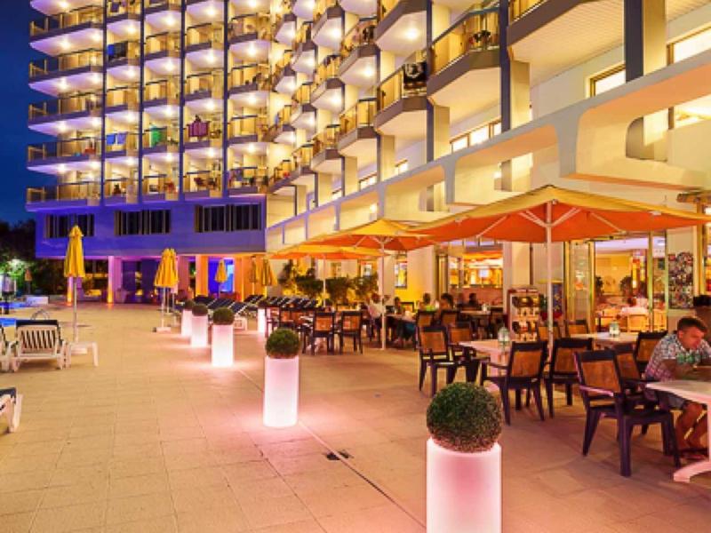 Osvětlený hotel s balkónovými pokoji a venkovní restaurací večer.