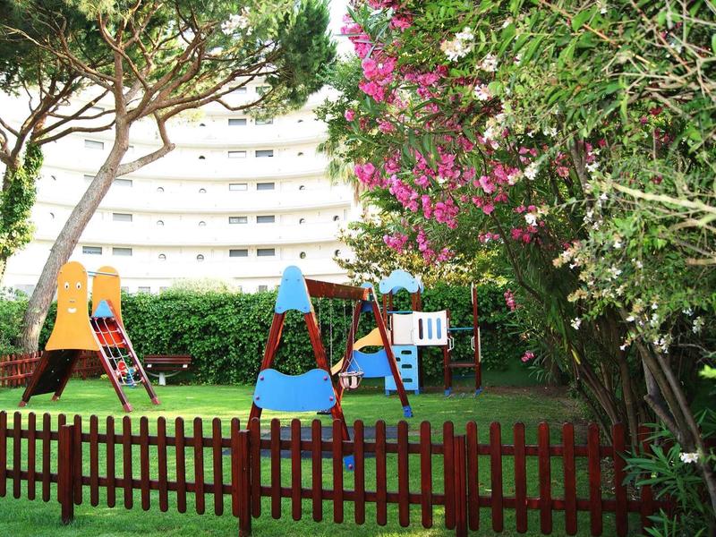 Dětské hřiště se skluzavkou a prolézačkou, obklopené stromy a keři poblíž budovy.