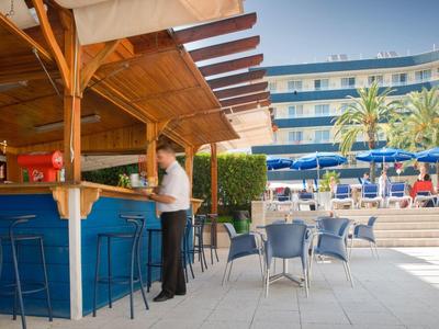 Bar al aire libre con techo de madera, taburetes azules y área de asientos junto a la piscina y hotel