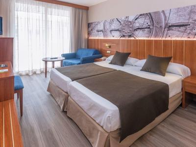 Habitación de hotel moderna con cama doble, sofá azul y ventana grande que deja entrar luz natural.