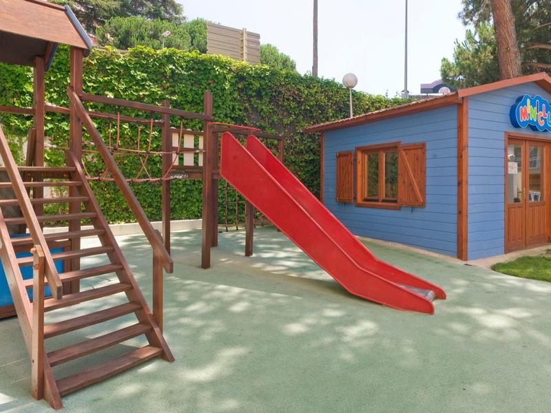 Área de juegos con tobogán, estructura para escalar y pequeña casita azul y roja en zona verde