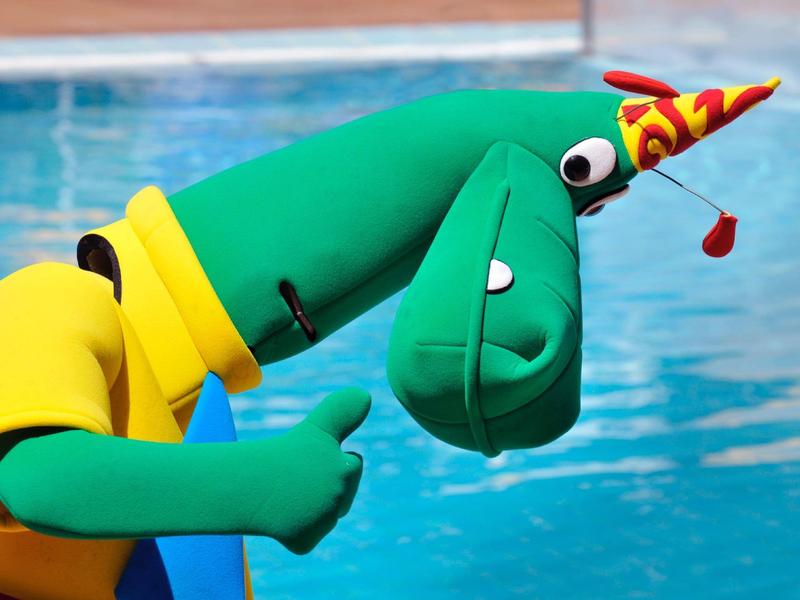 Figura inflable verde con sombrero de fiesta rojo y pulgar hacia arriba frente al agua de la piscina.