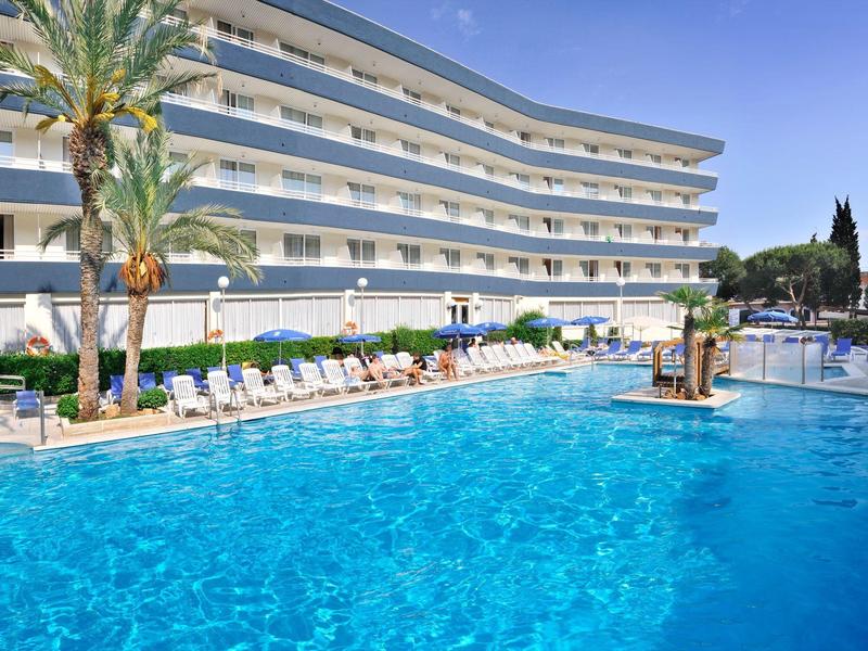 Gran piscina exterior frente a un hotel de varias plantas con tumbonas y palmeras.