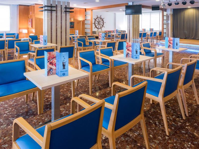 Gran sala de eventos con sillas azules y mesas blancas, escenario al fondo.