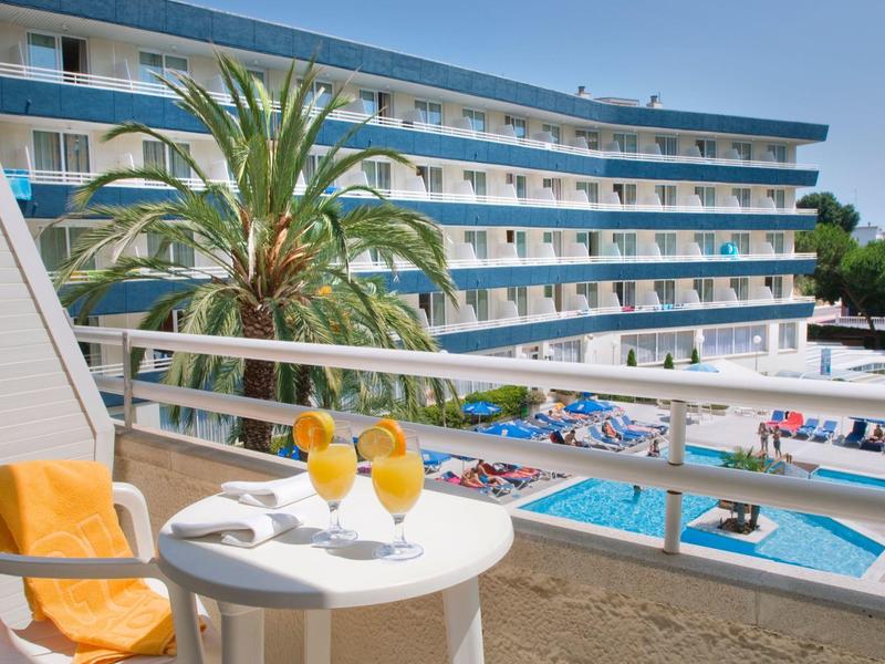 Vista de hotel con piscina y tumbonas, mesa con dos bebidas en balcón bajo el sol.