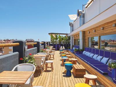Terraza de hotel con mesas y sillas de madera, cojines morados y vistas al cielo despejado.