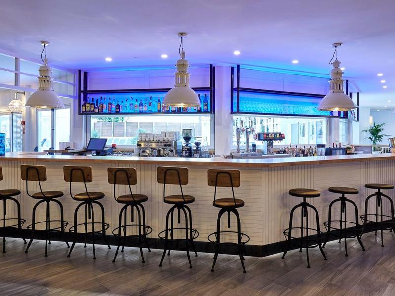 Área moderna de bar de hotel con mostrador iluminado y taburetes altos bajo lámparas colgantes.