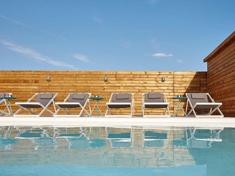 Área de piscina moderna con seis tumbonas grises y revestimiento de madera bajo un cielo despejado.