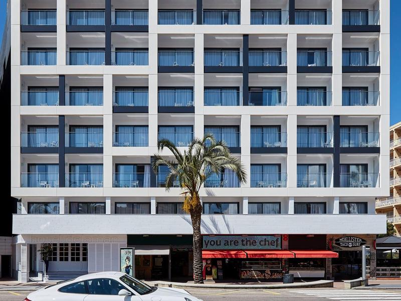 Edificio de hotel blanco moderno con balcones y un restaurante rojo en la planta baja.