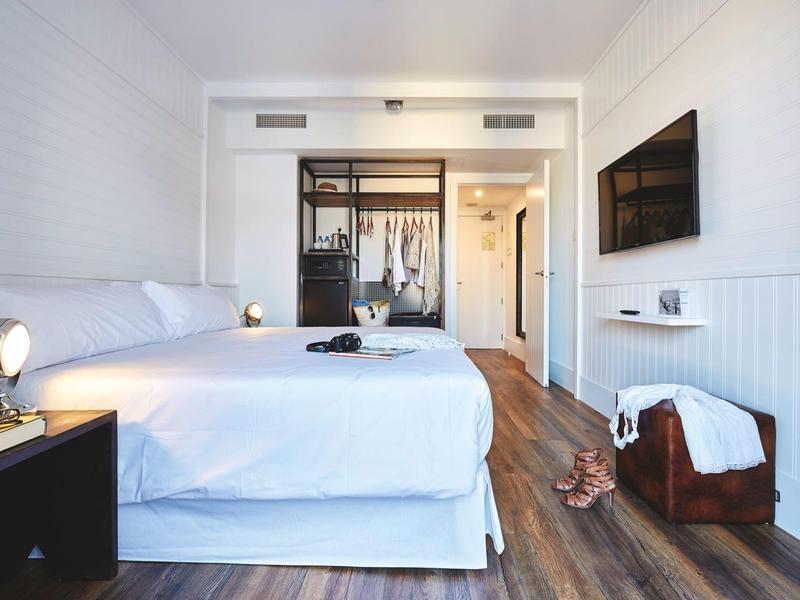 Habitación de hotel luminosa con cama doble, suelo de madera y decoración moderna, TV y armario abierto.