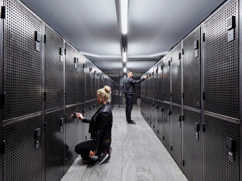 Sala de almacenamiento de equipaje en negro y gris con dos personas usando casilleros.