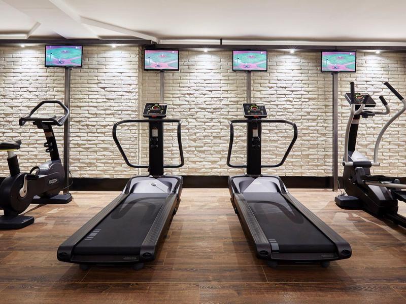 Gimnasio moderno con cintas de correr y máquinas elípticas frente a una pared de piedra blanca.