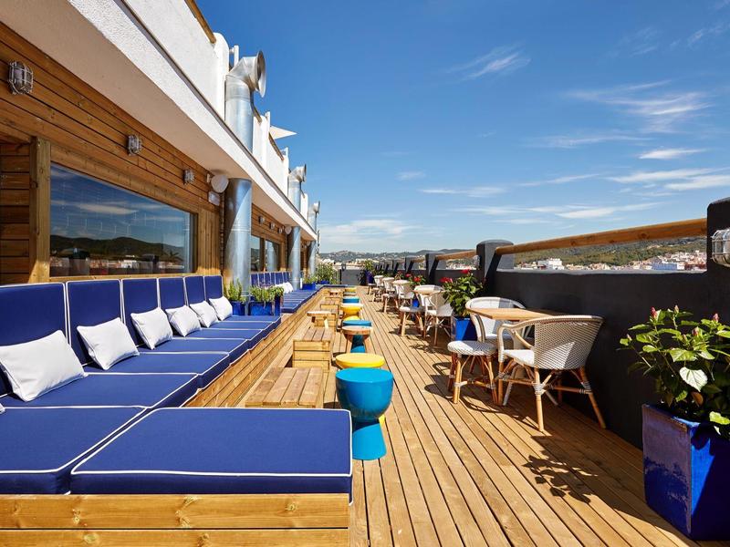 Terraza con tumbonas y mesas sobre un suelo de madera con vistas panorámicas y cielo azul.