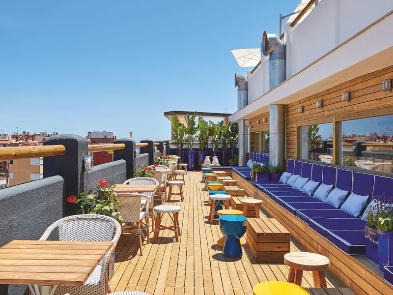 Terraza de hotel con mesas y sillas de madera, cojines morados y vistas al cielo despejado.