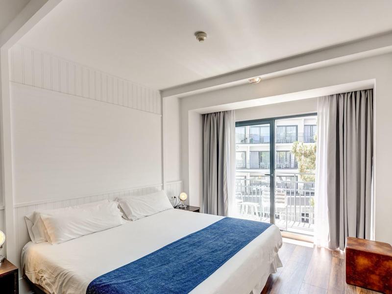 Habitación de hotel luminosa con cama grande, cubrecama azul, balcón y muebles modernos.