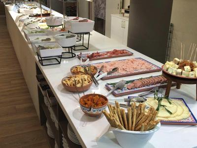 Long buffet with various foods like bread, cold cuts, and cheese in a modern room.