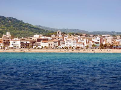 Coastal town with sandy beach, blue sea, and buildings against green hills under clear sky.