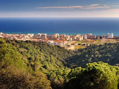 Coastal city with buildings near the sea, surrounded by green hills under a partly cloudy sky.