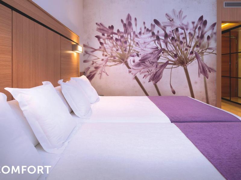 Modern hotel room with a large bed, white linens, purple accents, and floral wall art.