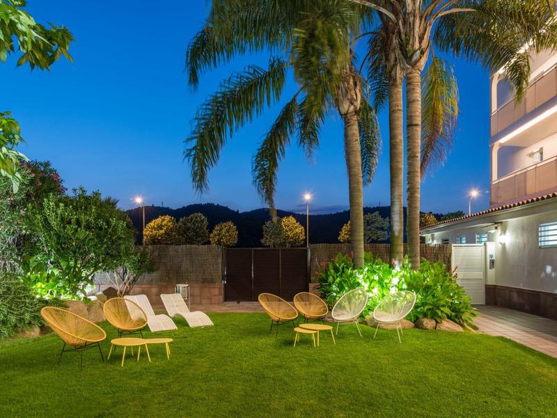 Night view of a garden area with wicker chairs, palm trees, and modern building lights.