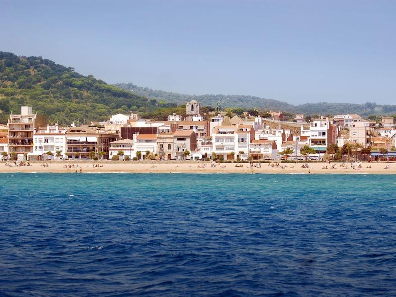 Coastal town with sandy beach, blue sea, and buildings against green hills under clear sky.