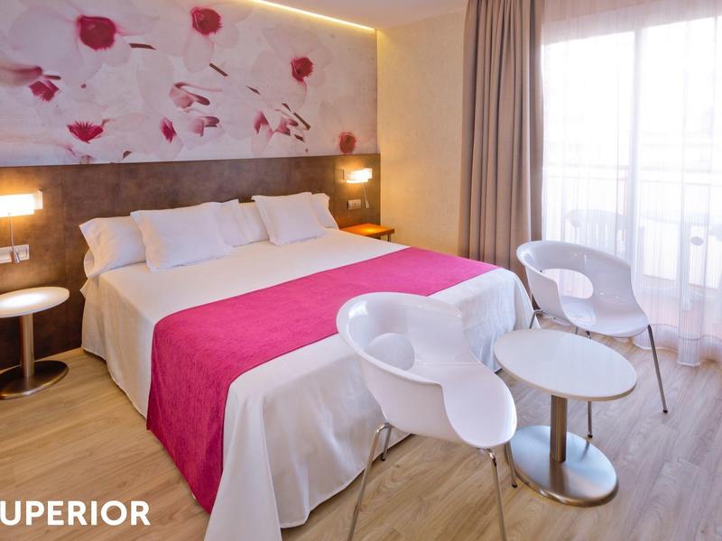 Bright hotel room with large bed, pink accents, modern white chairs, and sheer curtains.