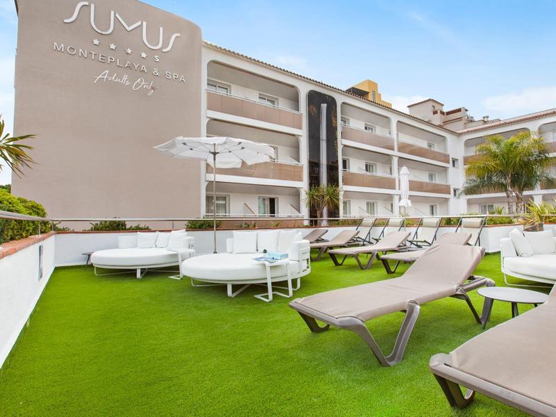 Sunlit hotel outdoor lounge with white sofas, sunbeds on green artificial grass under blue sky.