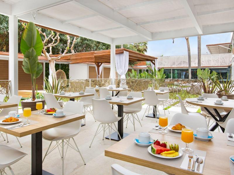 Outdoor hotel dining area with white tables and chairs, set with breakfast items and tropical plants.