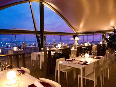 Elegancka restauracja z białymi stołami i krzesłami, oświetlona o zachodzie słońca z widokiem na morze.