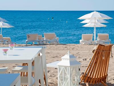 Plaża z białymi parasolami, leżakami i stołami z krzesłami na pierwszym planie.