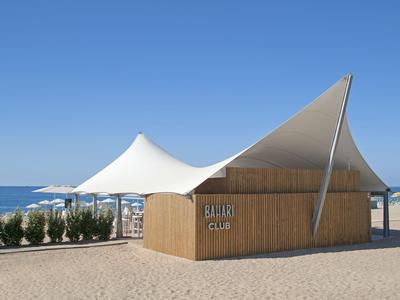 Modernes Strandclubgebäude mit weißem Zeltdach und Holzverkleidung an einem sonnigen Strand.