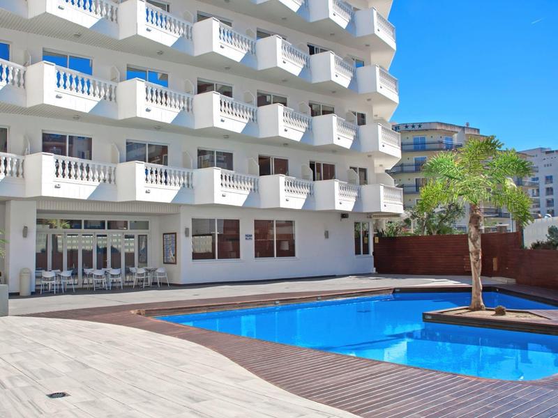 Nowoczesny basen hotelowy z drewnianym tarasem i palmami przed białym, wielopiętrowym budynkiem hotelowym.