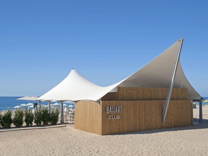 Nowoczesny klub plażowy z białym zadaszeniem namiotowym i drewnianą elewacją na piaszczystej plaży pod czystym niebem.