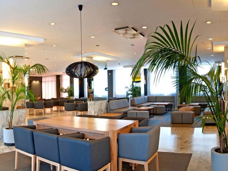 Jasny hol hotelowy z niebieskimi fotelami, drewnianymi stolikami i zielonymi roślinami.