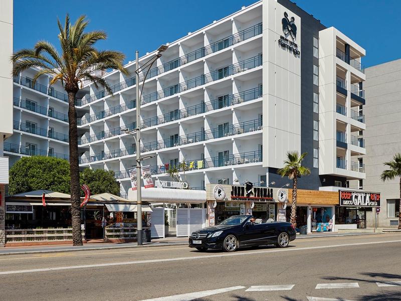 Edificio alberghiero moderno di sei piani con balconi e palme in una strada soleggiata.