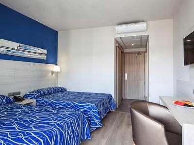 Camera d'albergo con due letti, biancheria blu a motivi, scrivania e porta verso il corridoio.