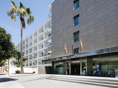 Ingresso di un hotel moderno con facciata in vetro e bandiere sotto un cielo soleggiato.