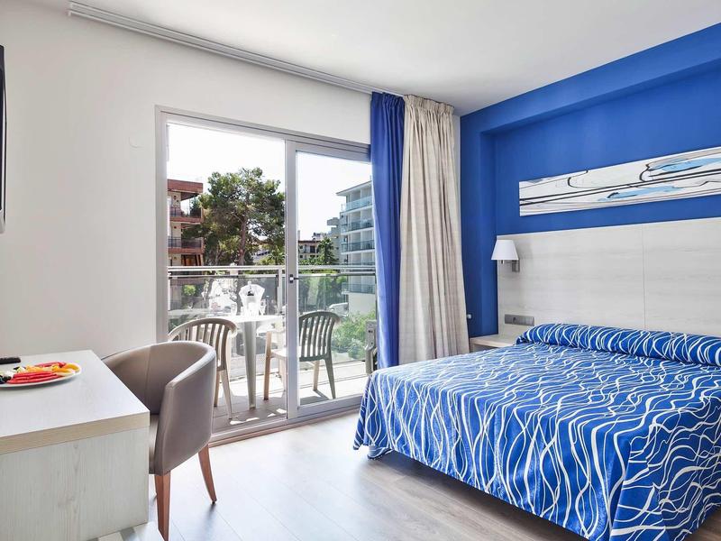 Camera d'hotel con accenti blu, letto matrimoniale e balcone con vista su giardino e edifici.
