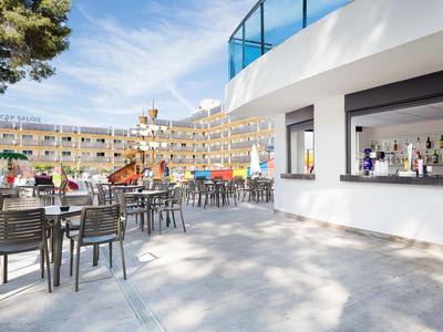 Outdoor hotel terrace with tables, chairs, and a bar under a bright sky.