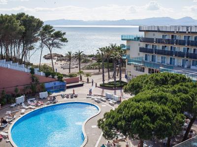 Hotel pool area with sun loungers, palm trees, and a view of the sea and distant mountains.