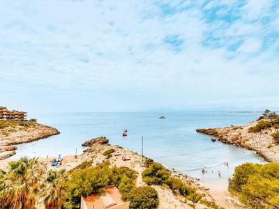 Coastal inlet with clear blue water, sandy beach, and scattered people under a partly cloudy sky.
