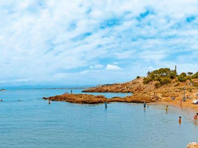 Sunny beach with calm sea, rocky outcrops, and buildings near the shore under a blue sky.