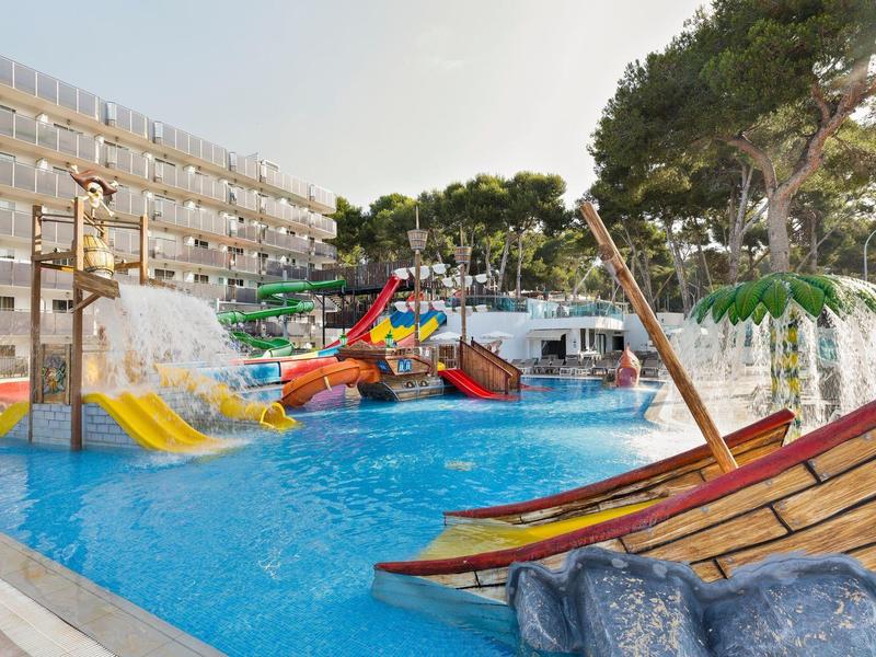 Outdoor hotel pool with water slides and pirate ship play area surrounded by sunlit trees.