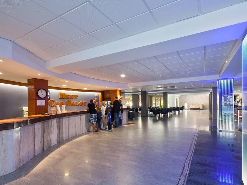 Spacious hotel lobby with reception desk, people, modern lighting, and tiled floor.