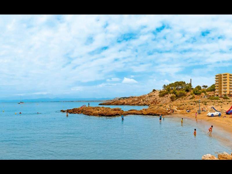 Sunny beach with calm sea, rocky outcrops, and buildings near the shore under a blue sky.