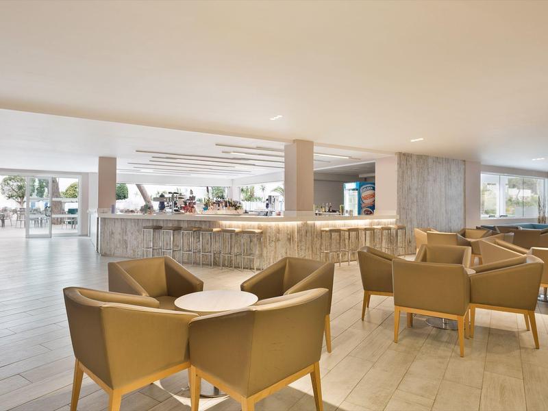 Modern hotel lobby with beige armchairs, wooden tables, and large windows with outdoor views.