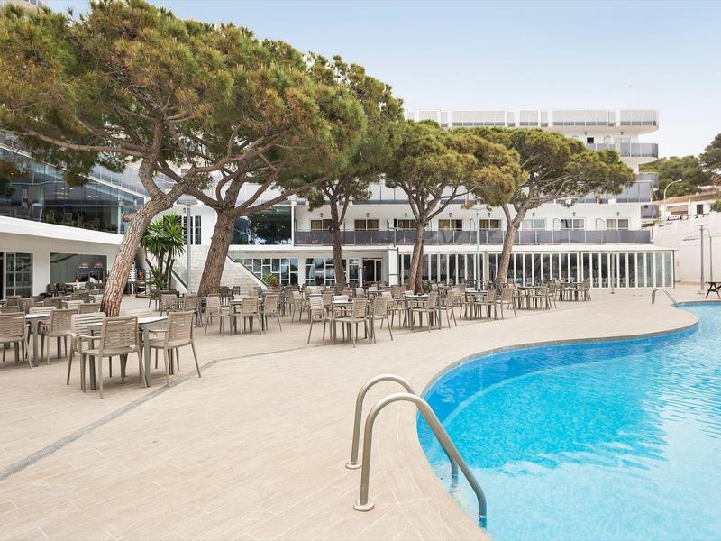 Empty outdoor pool area with blue water and beige chairs under green trees.