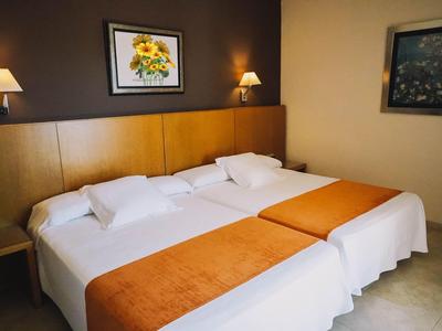 Dwa podwójne łóżka z białą pościelą i pomarańczowymi kocami w prosto urządzonym pokoju hotelowym.