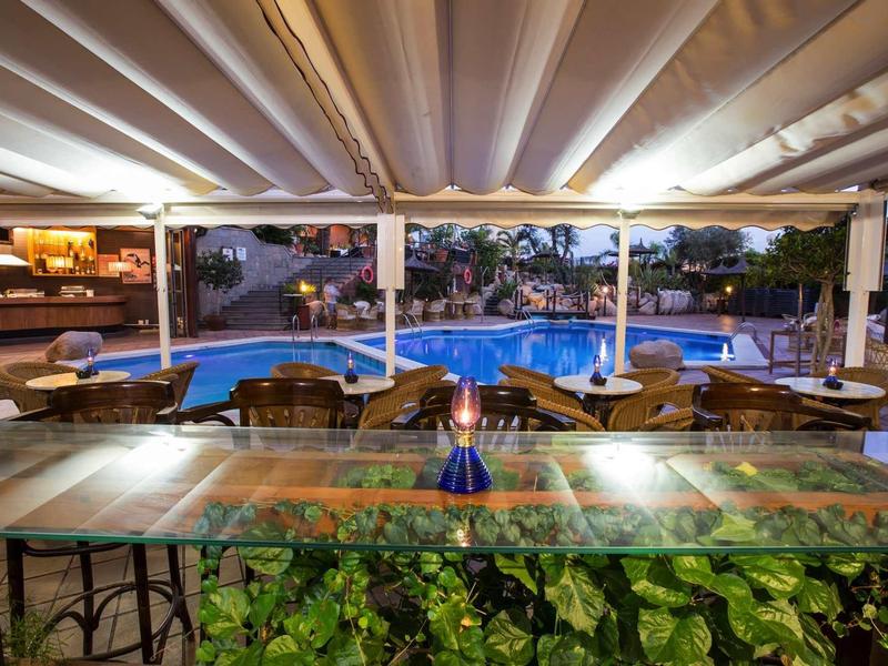 Outdoor hotel dining area with tables, chairs, and a lit fountain by a swimming pool at dusk.
