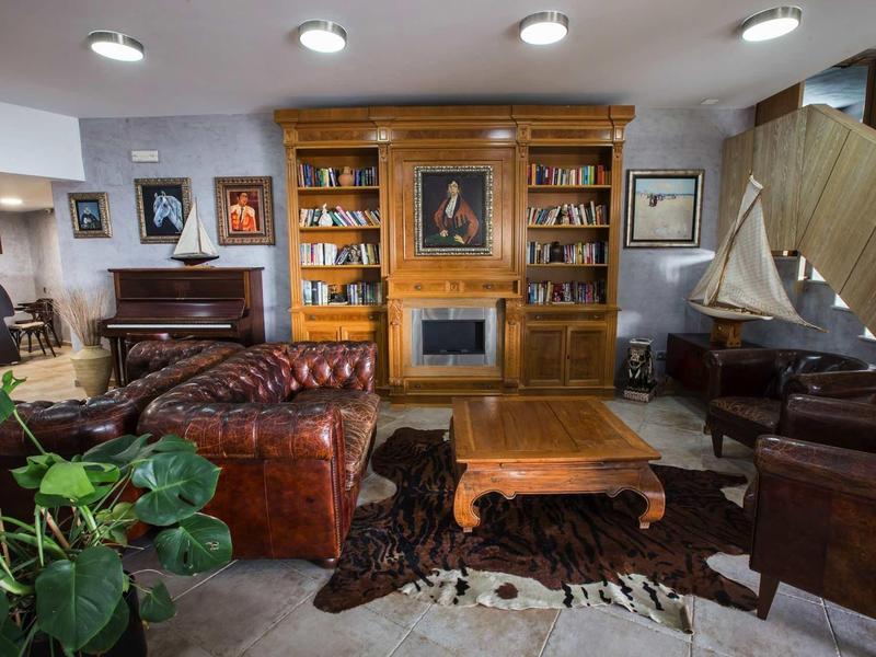 Cozy lounge with leather sofas, wooden bookshelf, coffee table, and indoor plants in a warm room.